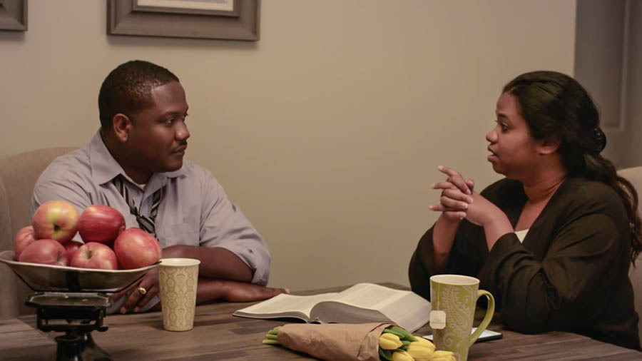 Hoton da aka dauko daga bidiyon nan ‘Wajibi ne Ku Yi Tsere da Jimiri’​—Ku Bi Dokokin Gasar.’ Dan’uwa da ’Yar’uwa Calou suna tattauna matsalolin da suke fuskanta cikin natsuwa. Sun kuma bude Littafi Mai Tsarki sun ajiye shi a kan teburi.