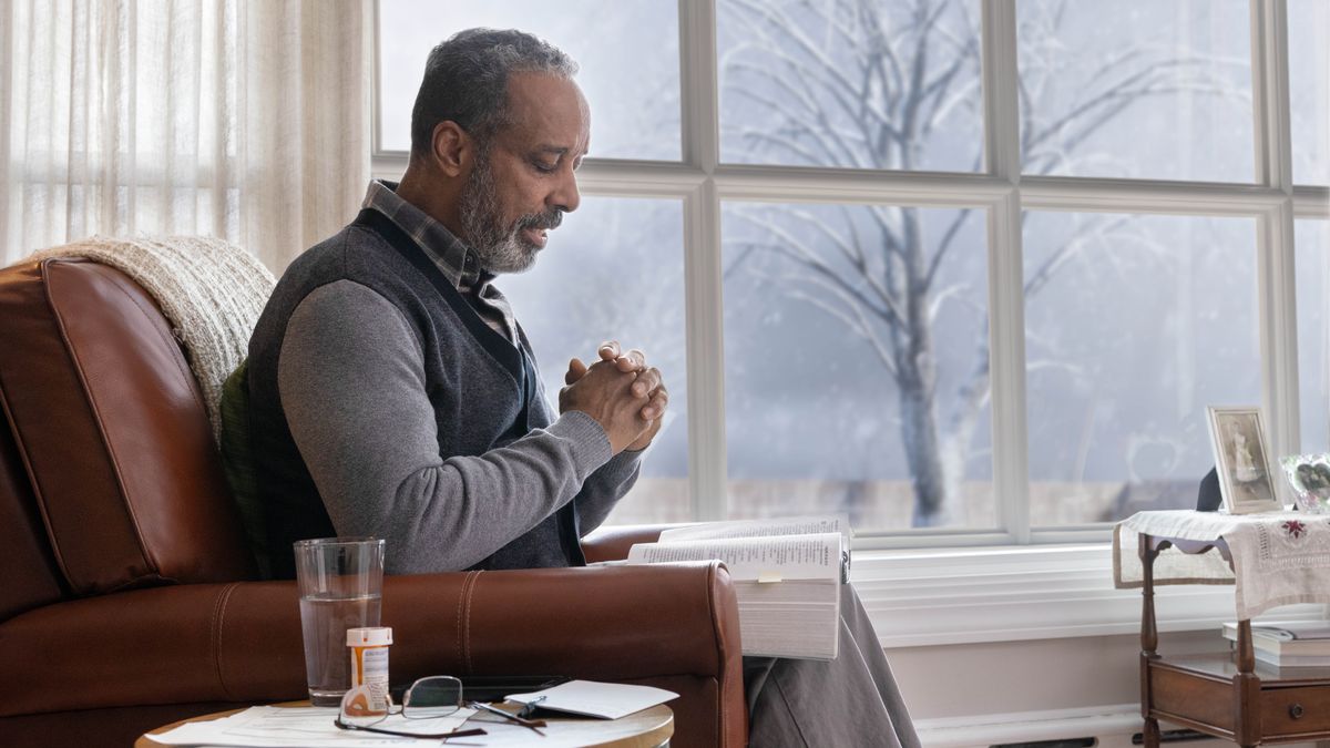 One olda braddah sitting at home an praying wintertime. He get one Bible wit him an one bottle of medicine on da table.