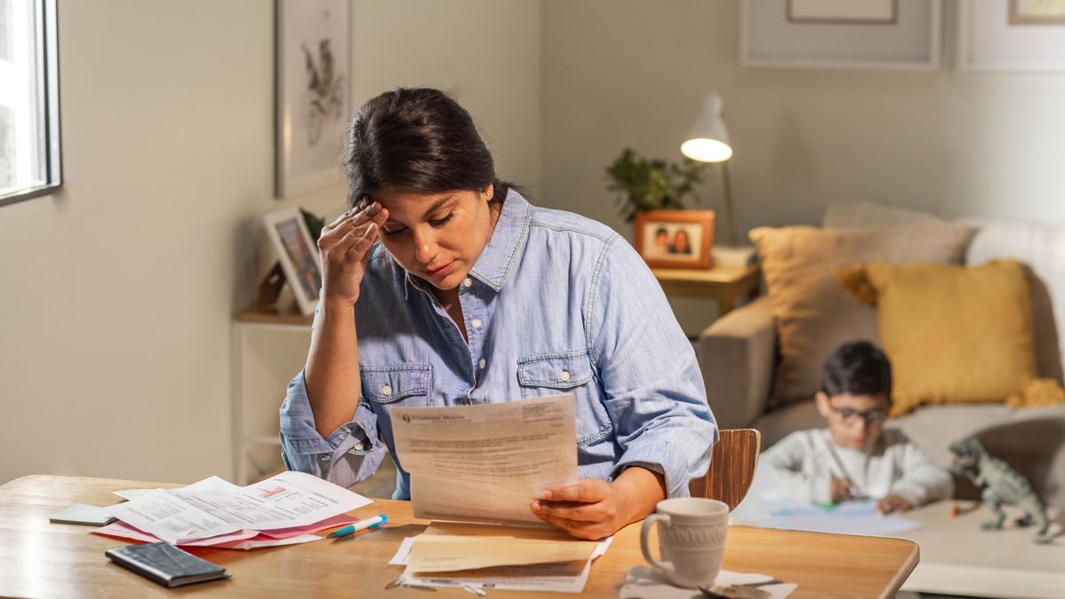 Ibu itu khawatir melihat tumpukan tagihannya, dan anak laki-lakinya bermain di belakangnya.