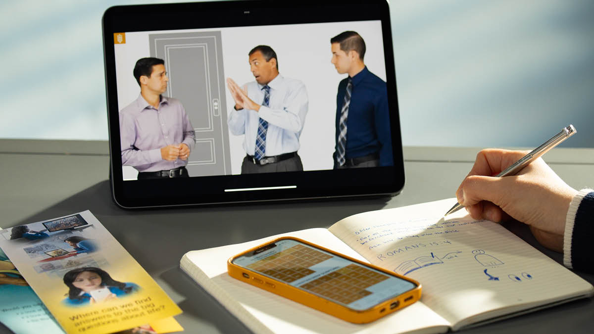 Una sorella prende appunti mentre guarda il video di una conversazione modello dell’adunanza infrasettimanale. Accanto al bloc-notes ci sono alcuni volantini e il suo telefono su cui è aperta l’app JW Library.