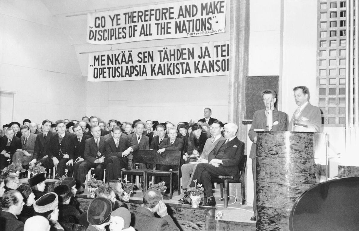 Fratelli e sorelle ascoltano il fratello Knorr mentre pronuncia un discorso avvalendosi dell’aiuto di un interprete. La scrittura dell’anno 1945 in inglese e in finlandese è appesa a un muro.