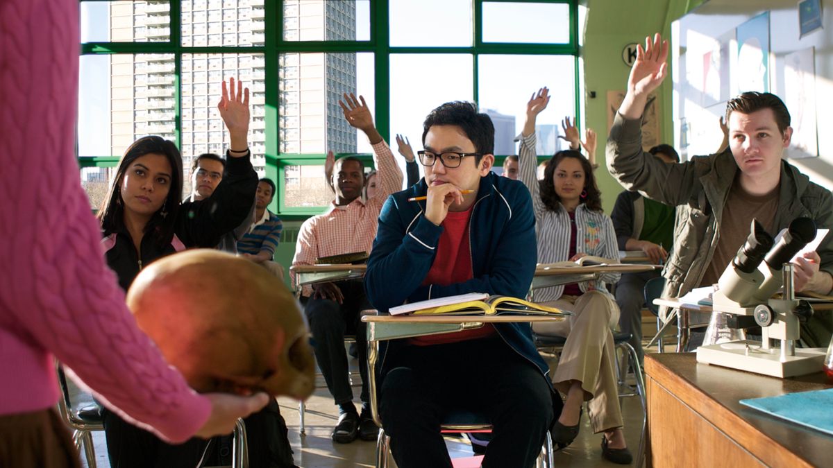 Un ragazzo a scuola riflette mentre osserva l’insegnante che tiene in mano un teschio dalla forma piuttosto insolita per essere quello di un essere umano. Mentre l’insegnante mostra il teschio alla classe, quasi tutti gli studenti hanno la mano alzata.
