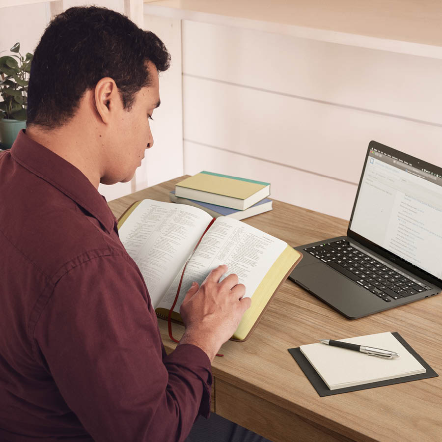 Wong lanang nggolèk keterangan ing Alkitab lan liwat laptopé. Ing méja ya ana buku cathetan.