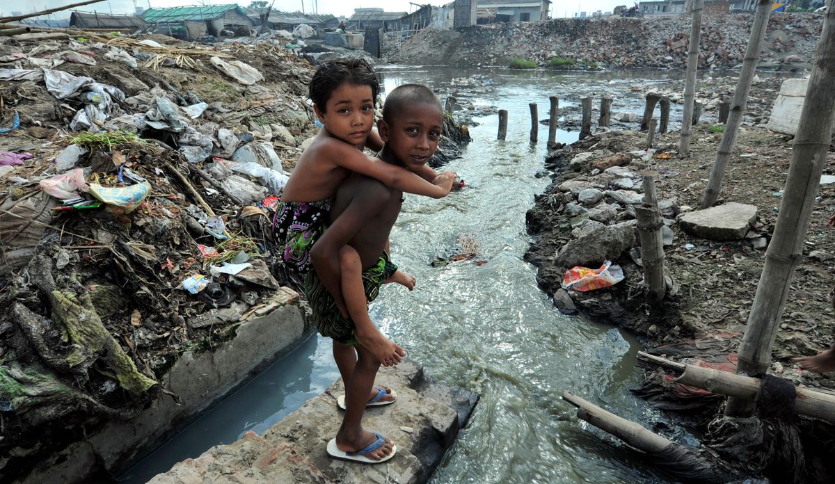 Cah lanang nggéndhong adhiné. Ing sekitaré ana akèh sampah lan banyu sing tercemar.
