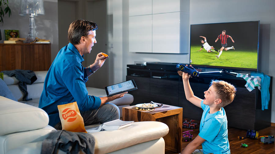 Sedulur lanang sing sinau Alkitab disambi mangan lan nonton TV. Ing sebelahé, anaké lagi dolanan.