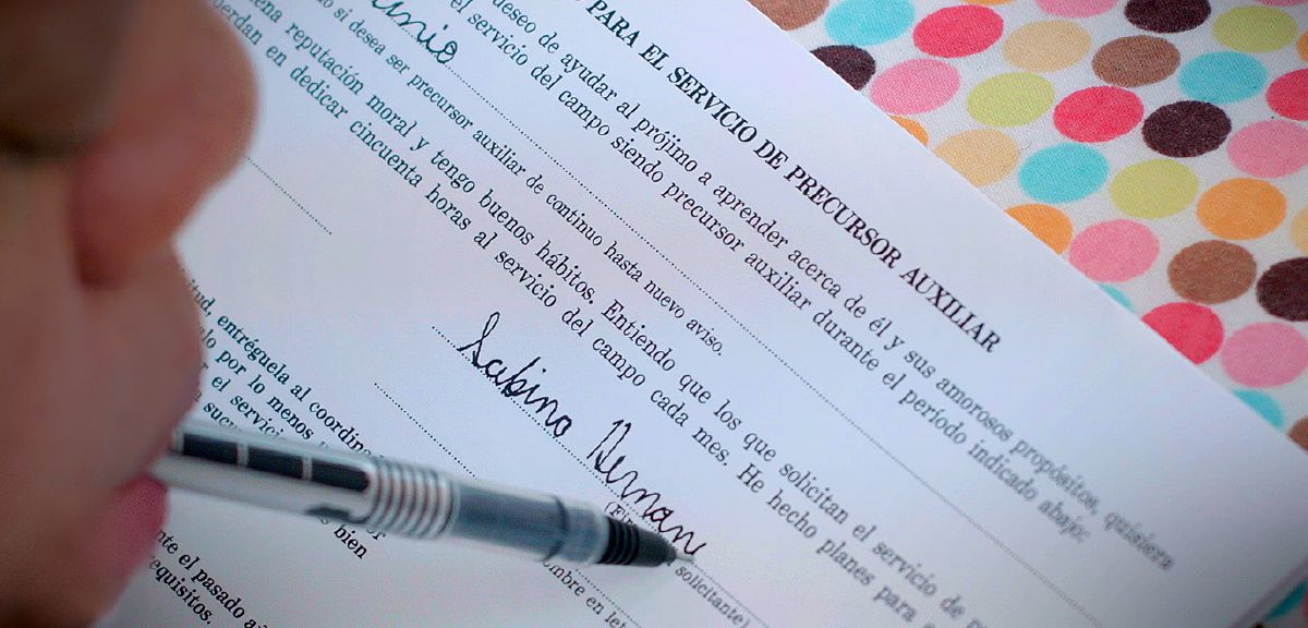 საბინა ჰერნანდესს კბილებით უჭირავს კალამი და ხელს აწერს დამხმარე პიონერობის ბლანკს