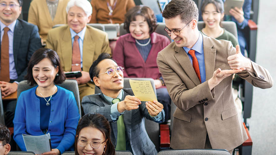 외국어를 배우는 형제가 왕국 회관에서 열린 집회에서 쪽지에 적어 놓은 해설을 읽고 있습니다. 참석한 다른 사람들이 존중심 있게 그 모습을 바라봅니다.