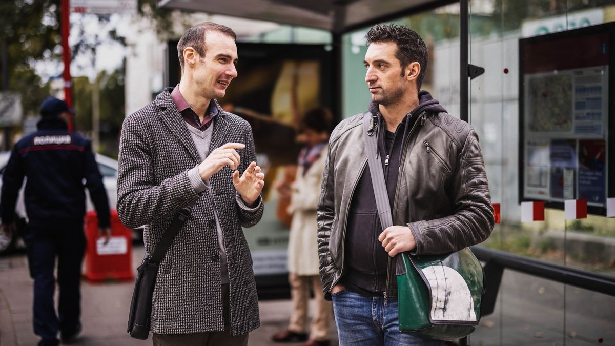 Ow’oluganda ayambadde engoye ez’ekisaazisaazi ng’abuulira omusajja mu kifo bbaasi we zisimba. Omusirikale abali mabega ng’adda ku luuyi olulala.