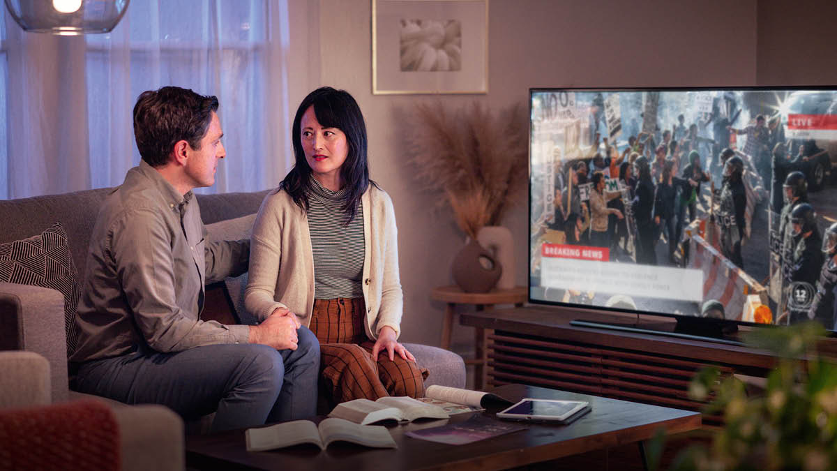 Un couple regarde les informations à la télévision ; il y a des troubles civils dans le pays. Une Bible et d’autres publications sont posées sur la table qui se trouve devant eux.