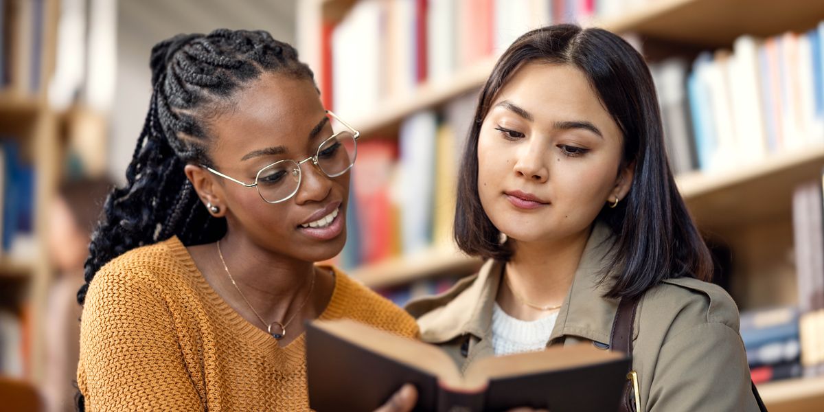 Две девојки заедно разгледуваат една книга во библиотека.