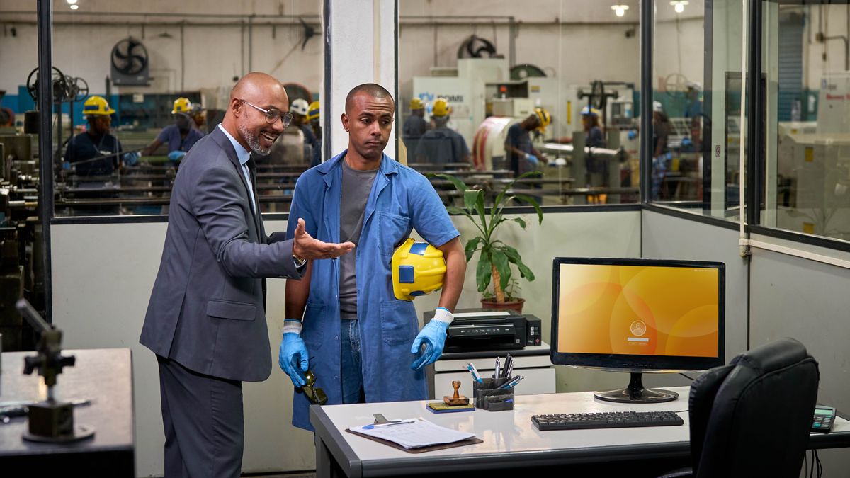 Um irmão com capacete e roupa de trabalho num escritório vazio junto com seu supervisor. O supervisor diz que a vaga de trabalho está disponível e oferece a ele uma promoção.