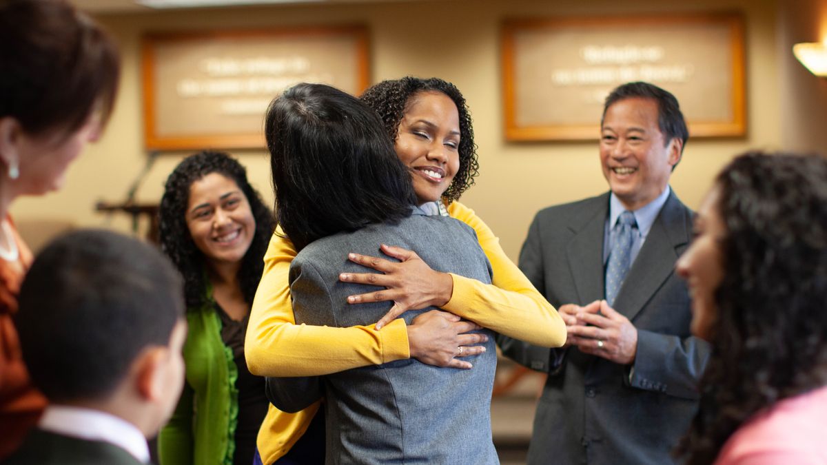 Two sisters hugging each other at the Kingdom Hall. Others happily gather around them.