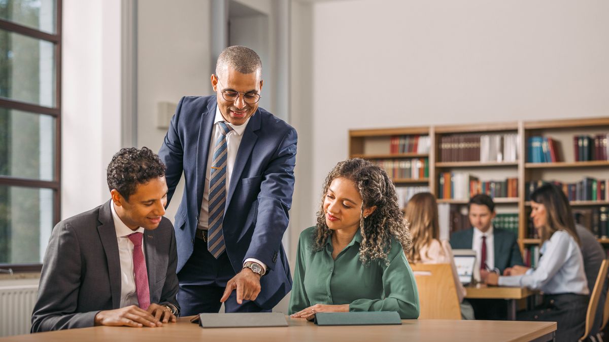 Um instrutor e um casal usando um dispositivo eletrônico para conversar sobre um assunto espiritual enquanto estudam em uma biblioteca na Escola para Evangelizadores do Reino. Dá para ver outros alunos estudando.