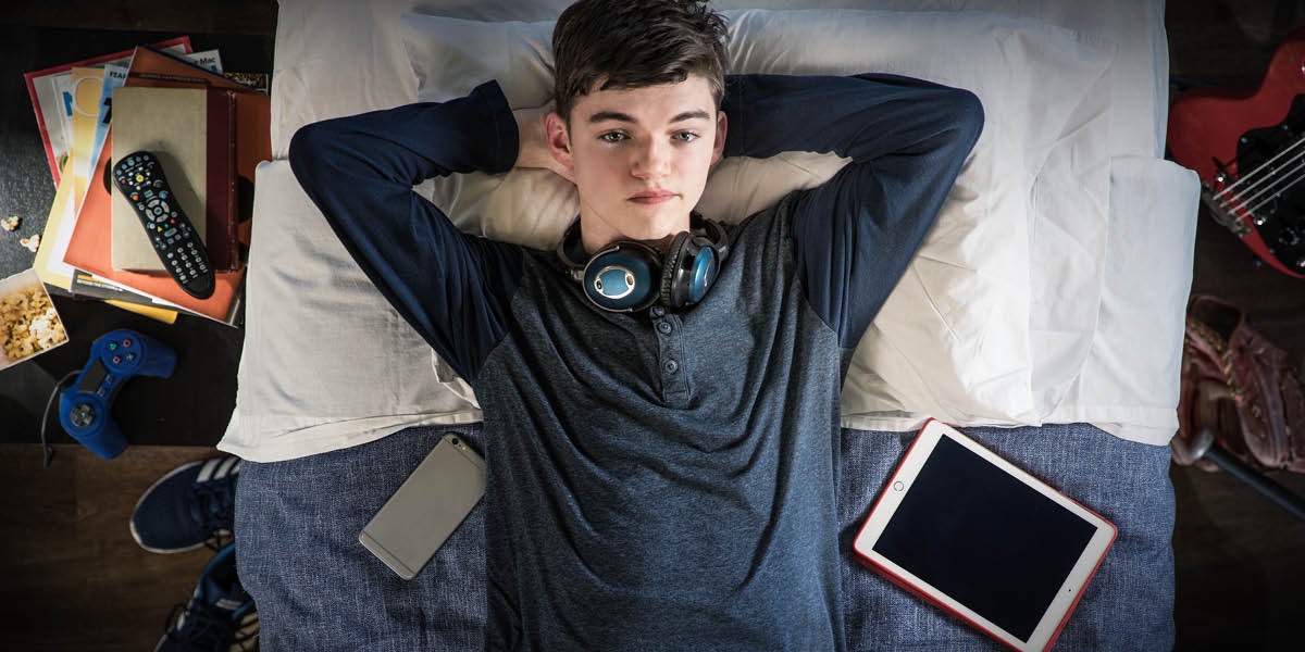 Um adolescente com muitos aparelhos eletrônicos, mas deitado na cama olhando para o teto