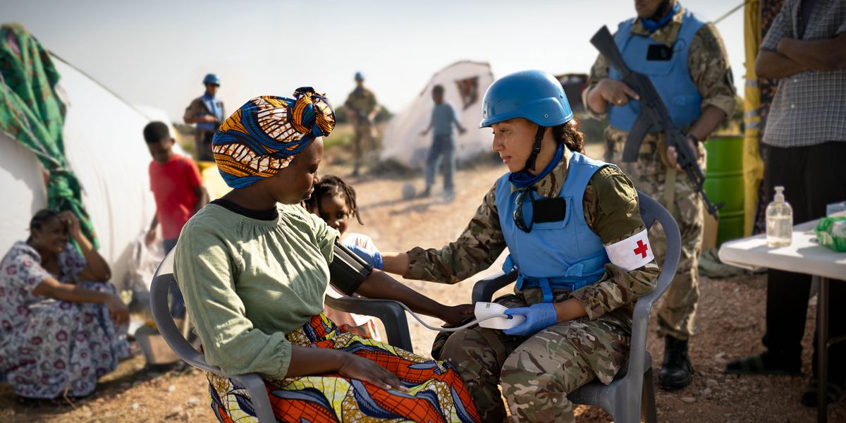 Uma médica conferindo os sinais vitais de uma refugiada. Soldados armados das forças de paz e outros refugiados estão ao redor.