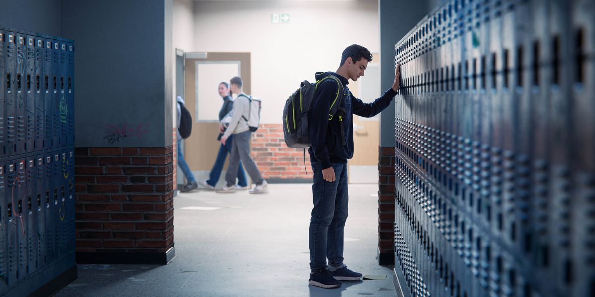 En el pasillo de una escuela, un adolescente triste está de pie frente a unos casilleros o taquillas.