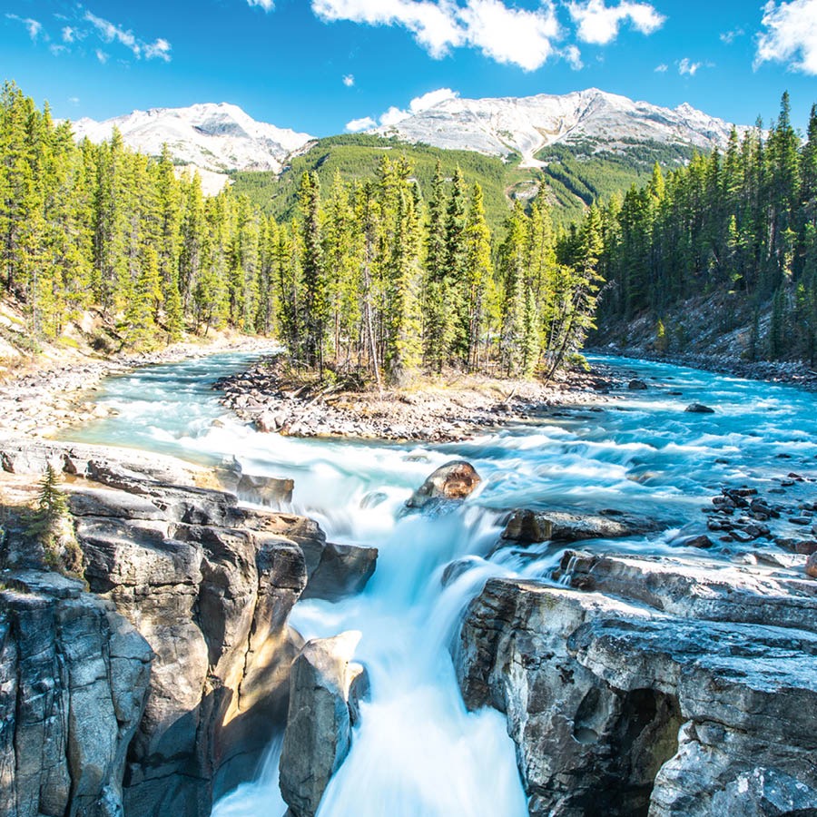 Un río que desemboca en una cascada. Está rodeado de montañas nevadas y de un bosque