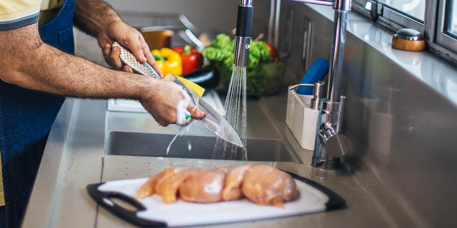 Мужчина моет нож водой с мылом. Сырое куриное мясо и овощи лежат на разных разделочных досках.
