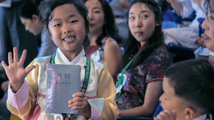A scene from the video ‘‘Love Never Fails’! International Conventions.’ A happy girl holds up her copy of the ‘New World Translation’ that she just received.