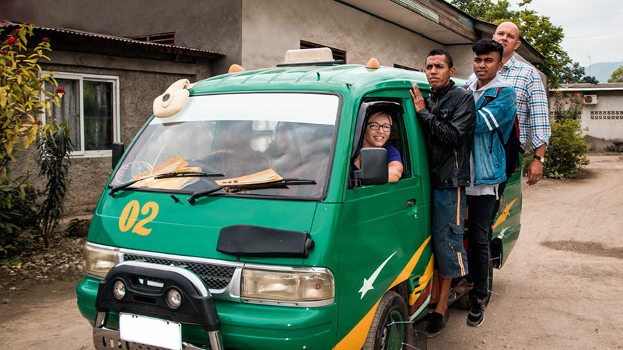 Vanessa, Alex, and others riding on a small bus that is packed with people and has three brothers hanging on the outside.