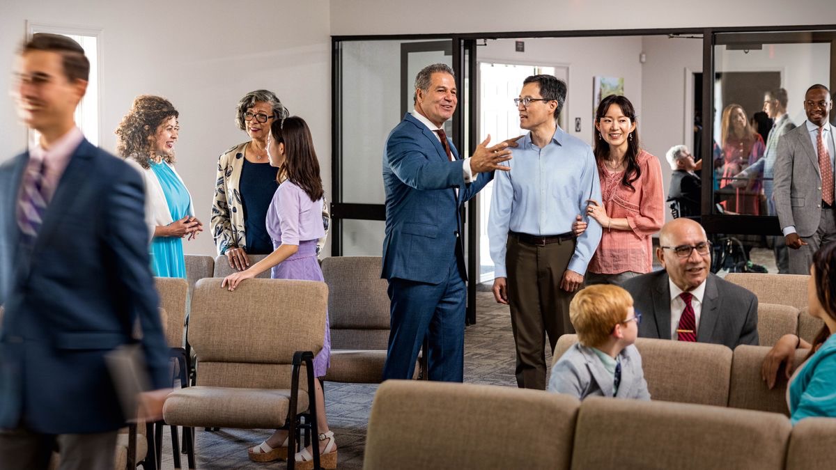 Brothers and sisters of different ages and backgrounds, happily conversing before a congregation meeting. A brother welcomes a non-Witnesses couple.