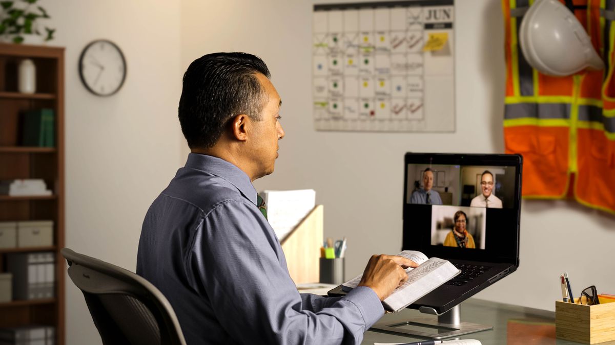 Two elders making a shepherding call on a sister via videoconferencing late at night. One of the elders reads from the Bible. Construction gear and a calendar filled in with many appointments hang on a wall in front of him.