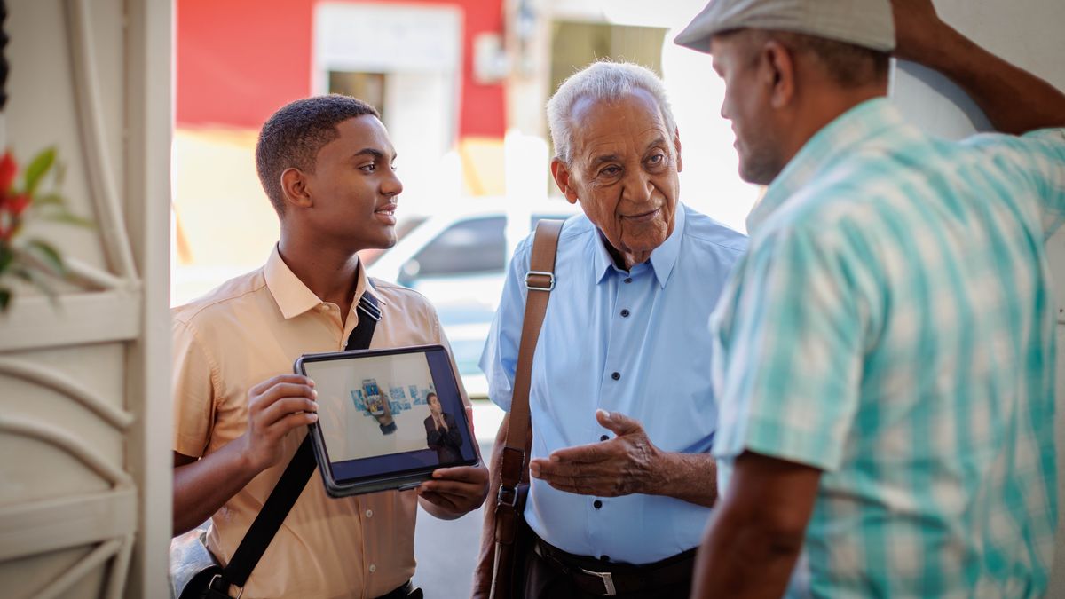 A young brother and an elderly brother working together in the house-to-house ministry. The elderly brother talks with a man while the young brother holds up a tablet that is playing a video.
