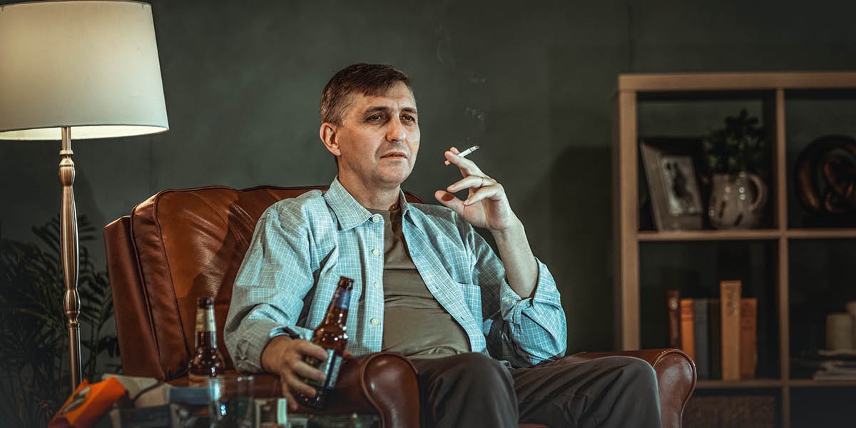 En man sitter i en fåtölj med en cigarett i ena handen och en ölflaska i den andra.