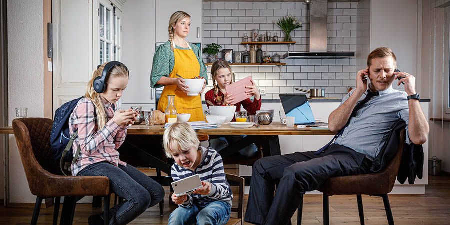 En mamma sätter fram frukost och ser irriterad ut när resten av familjen bara sitter med sina elektroniska enheter.