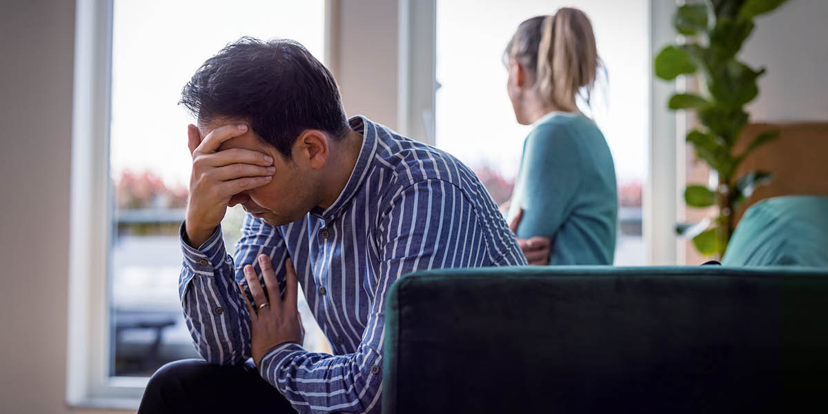 En ledsen man gömmer ansiktet i sina händer medan hans fru uppgivet tittar ut genom ett fönster.