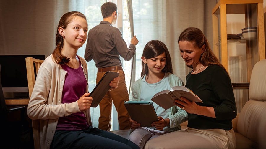 Brat sa doma pozerá cez okno a dáva pozor, zatiaľ čo si jeho manželka číta s dvomi dcérami Bibliu