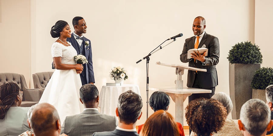 Bwana na bibi harusi wakifunga ndoa katika Jumba la Ufalme. Mhudumu wa Mashahidi wa Yehova akiendelea kutoa hotuba  inayotegemea Biblia.