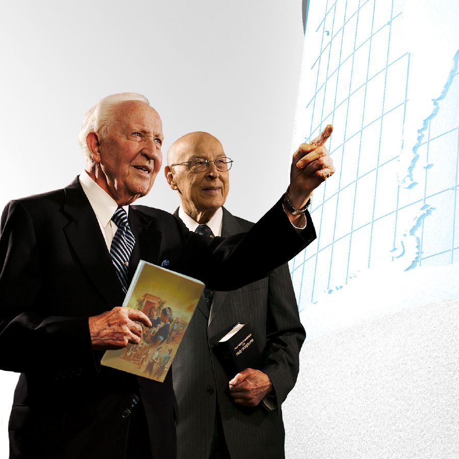 John Barr y Theodore Jaracz, miembros del Cuerpo Gobernante, viendo un mapa del mundo. El hermano Barr está sujetando el libro “Demos ‘un testimonio completo sobre el Reino de Dios’”.