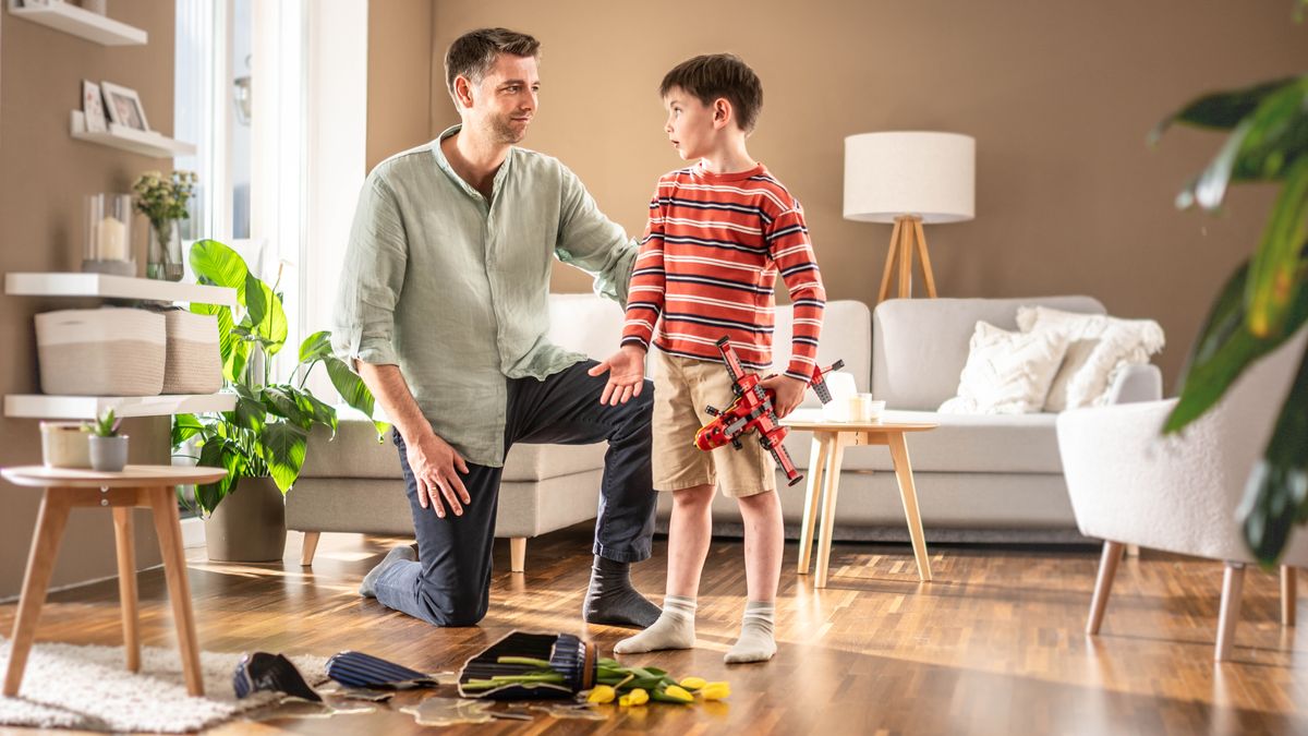 Un padre escucha con paciencia a su hijo, que le está explicando que rompió sin querer un jarrón con flores mientras jugaba con un avión de juguete.