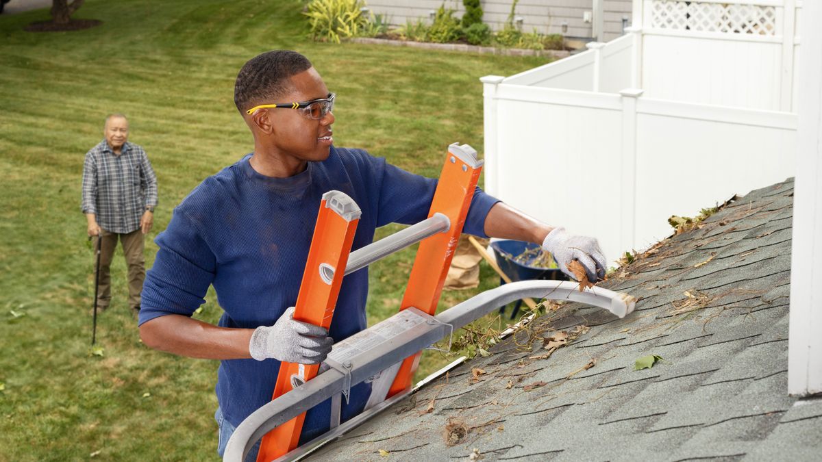 Un jeune frère est monté sur une échelle ; il retire les feuilles qui se trouvent dans une gouttière de la maison d’un frère âgé.