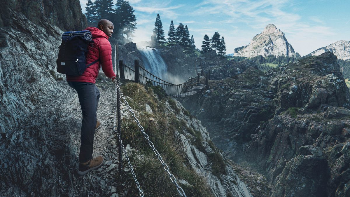 Umfana osemtsha obeboniswe kwinqaku elingaphambili uhamba kwindawo elithambeka nencinci enamatsheyina okubambelela. Phaa kude kubonakala iblorho nentaba.