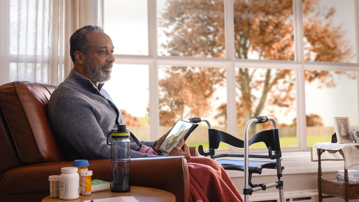 The elderly brother sitting in his home in the autumn season and thinking about the picture of Paradise displayed on his tablet. Beside him is a walker and several bottles of medication.