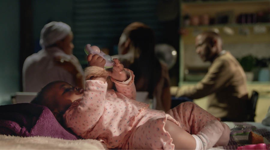 A baby lying on a bed, wearing an amulet on its wrist. In the background, the baby’s mother, a Bible student, is talking to the couple who studies the Bible with her.