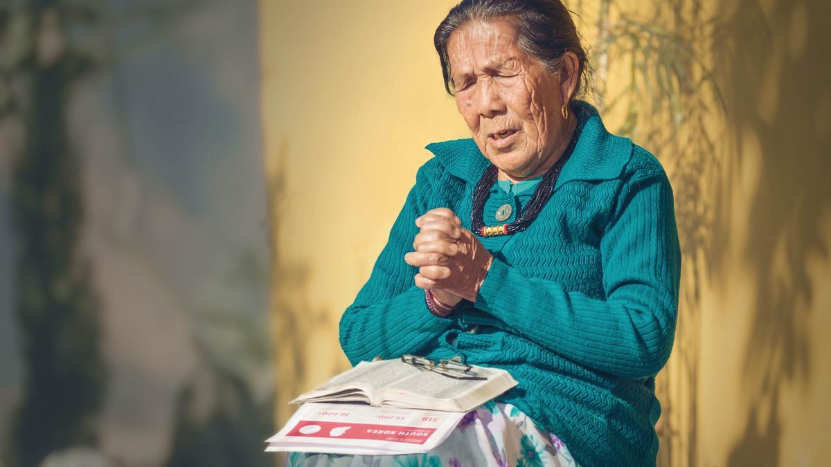 An elderly sister praying intensely. On her lap are a Bible and a list from jw.org of brothers in prison.